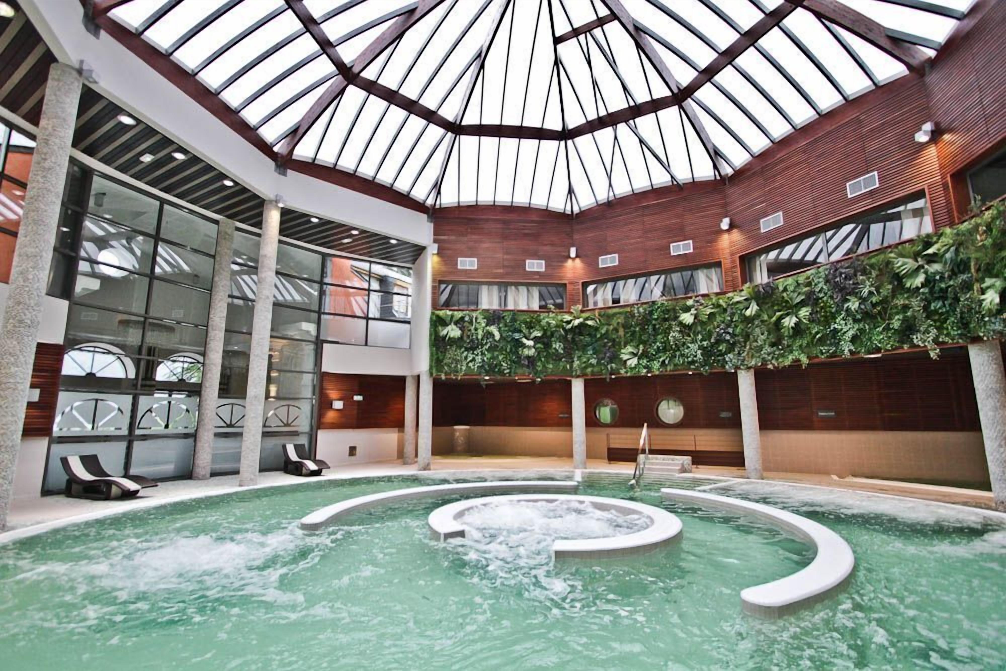An indoor swimming pool with a glass ceiling, surrounded by lounge chairs and a lush green wall.