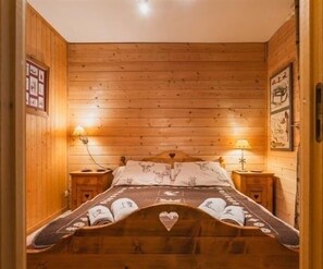 Une pièce aux murs lambrissés, avec un lit agrémenté d’une couvre-lit à motifs et d’un oreiller en forme de cœur. On y trouve deux lampes de chevet et un tableau encadré accroché au mur.