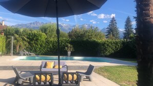A pool area with lounge chairs and an umbrella, surrounded by greenery and mountains in the background.
