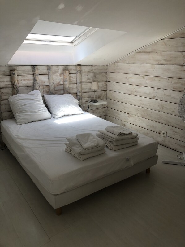 A bedroom with a skylight, a wooden headboard, a bed with white linens, a bedside table with a lamp, and a stack of towels. - Large house 9P + C 200m² with individual swimming pool, beautiful view of mountains (Crolles)
