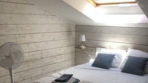 A bedroom with a skylight, a bed with pillows, a bedside table, a lamp, and a standing fan.
