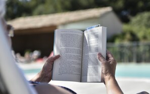 Eine Person hält ein offenes Buch mit sichtbarem Text in der Hand, draußen in der Nähe eines Schwimmbeckens.