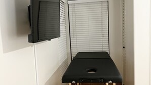 Een kamer met een zwarte massagetafel, een wandgemonteerde televisie en een raam met jaloezieën.