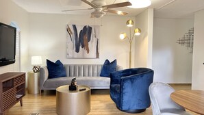 Een moderne woonkamer met een grijze bank, een blauwe fauteuil, een houten salontafel, een vloerlamp en een plafondventilator.