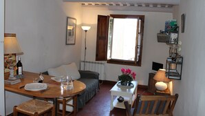 A cozy room with a bed, a dining table with chairs, a small white table with a lamp, and a window with curtains.