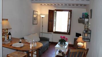 Une pièce confortable avec un lit, une table à manger entourée de chaises, une petite table blanche avec une lampe et une fenêtre avec des rideaux.