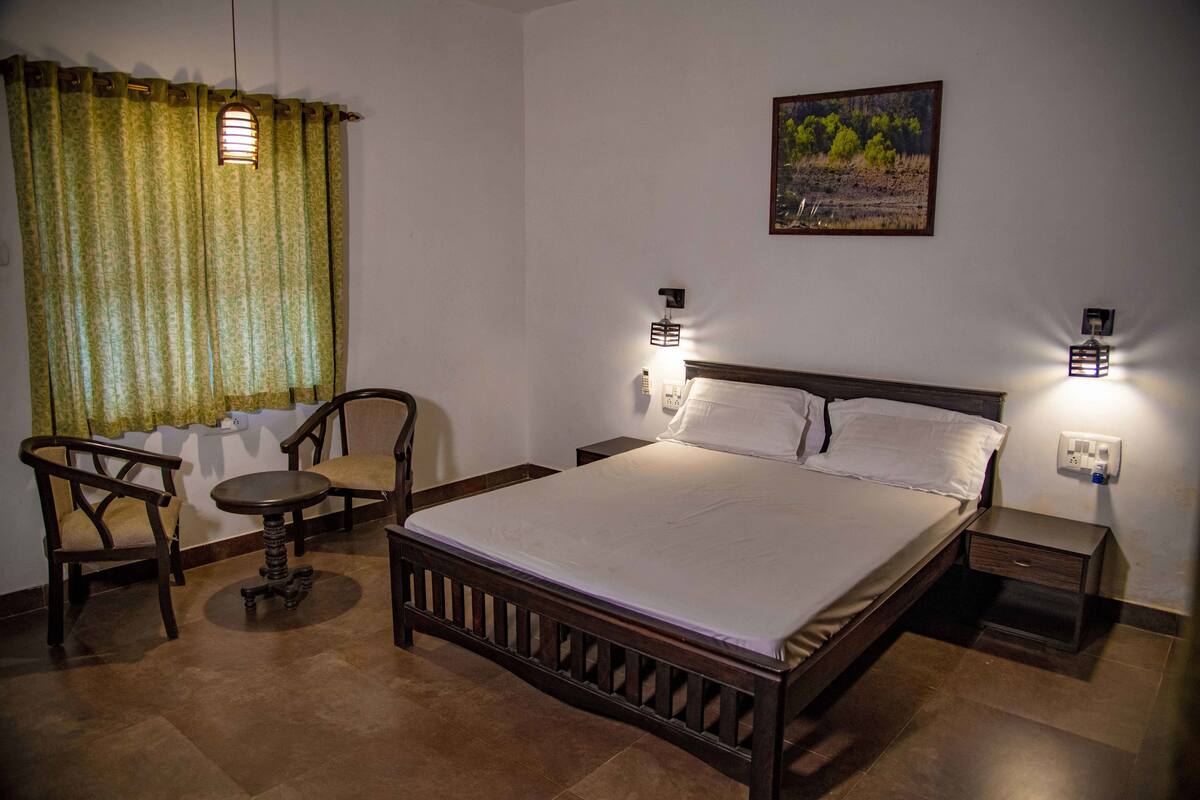 Quarto com cama de madeira, duas mesas de cabeceira, um criado-mudo, uma cadeira, uma mesa redonda pequena e uma janela com cortinas.