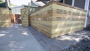 Una estructura de madera con listones horizontales, posiblemente un almacenamiento o separador, junto a un edificio con cimientos de piedra y una valla metálica al fondo.