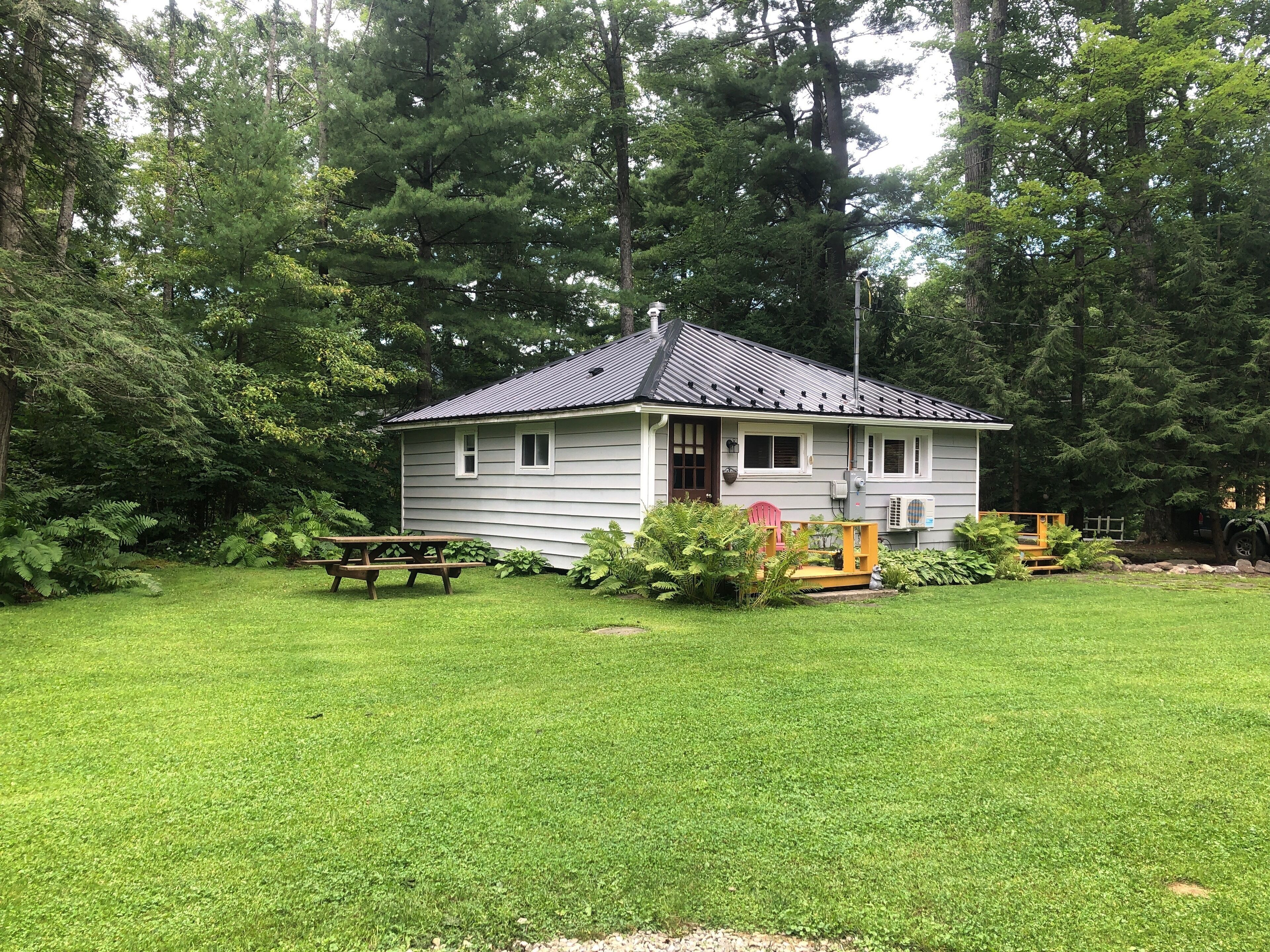 A small, single-story cabin with a dark roof, surrounded by a well-maintained lawn and dense forest.