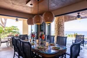 Un coin repas avec une table dressée pour quatre personnes, entourée de chaises en osier, et offrant une vue sur l’océan.