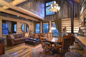 L’intérieur d’une cabane en rondins, avec un plafond en bois, de grandes fenêtres, un lustre, des meubles en cuir et une cheminée.