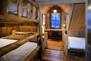 A rustic cabin interior with wooden walls, a desk, two beds, and a window with a view.