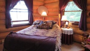 Un dormitorio de cabaña de troncos con una cama grande, dos ventanas con cortinas, una mesa redonda con una lámpara y una silla pequeña.