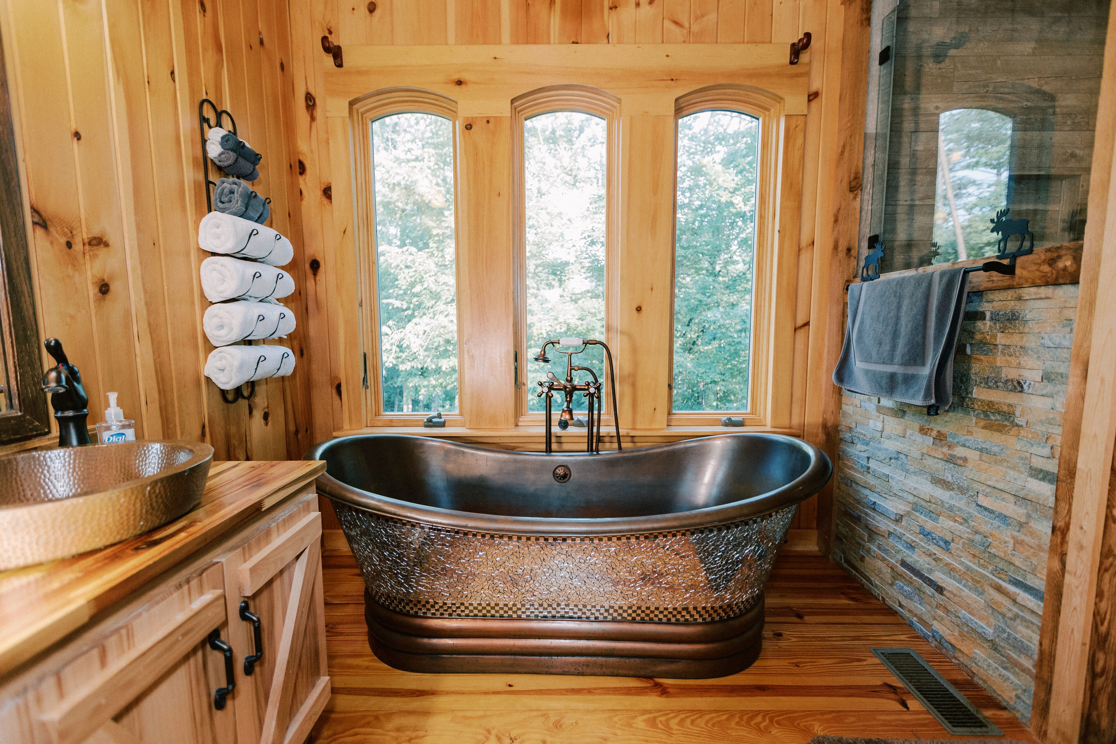 A wooden cabin bathroom with a freestanding bathtub, a wooden vanity with a sink, and a window with a view of trees.