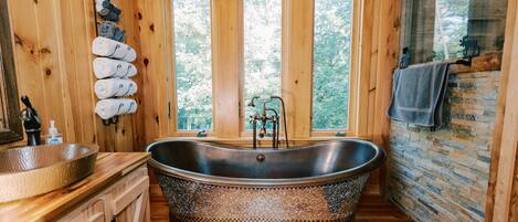 A wooden cabin bathroom with a freestanding bathtub, a wooden vanity with a sink, and a window with a view of trees.