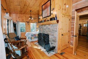 Interior acogedor de una cabaña con chimenea de piedra, paredes de madera, una cama con colcha floral, una mecedora y un comedor.