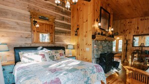 Ein gemütliches Schlafzimmer in einer Blockhütte mit einem großen Bett, einem Kamin aus Stein, Holzwänden und einem angebrachten Fernseher.