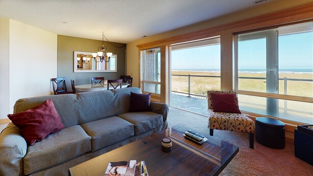 A living room with a sofa, armchairs, a coffee table, and a dining area with chairs and a table. Large windows offer a view of the beach.