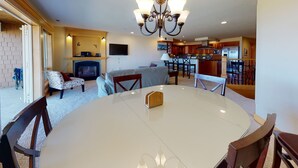 A spacious living room with a fireplace, a dining area with a round table, and a kitchen in the background.