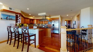 A spacious kitchen with wooden cabinets, a central island with bar stools, a stainless steel refrigerator, and a dining area with a black wrought iron railing.