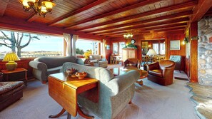 A cozy, well-lit living room with wooden beams, comfortable seating, a dining area, and a view of the outdoors.