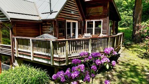 A wooden cabin with a deck, surrounded by lush greenery and purple flowers. - Fayette Springs--Creek House--Enjoy this Spacious Cottage! (Farmington)