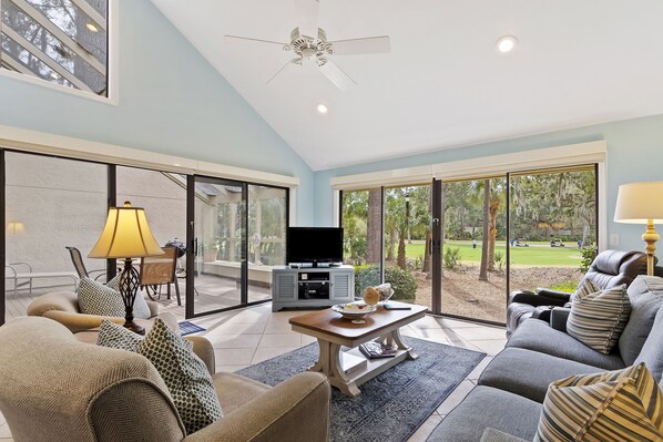 A spacious living room with a sofa, armchair, and a coffee table. There is a television and a cabinet in the background.