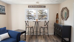 Une pièce avec un canapé bleu, deux fauteuils, une petite table sur laquelle se trouvent une bouteille et un verre, un meuble et une fenêtre donnant sur des arbres.
