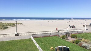Vista de la playa con una cerca blanca, una zona verde y una torre de salvavidas a lo lejos.
