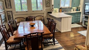 Een eethoek met een houten tafel en stoelen, een keuken met blauwgroene kastjes en een raam met jaloezieën.