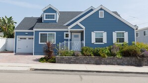 Une maison bleue à deux étages, avec une porte de garage blanche, un petit porche à l’avant et un mur de soutènement en pierre agrémenté de plantes.