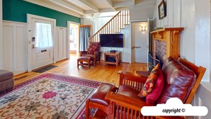 A spacious living room with a wooden floor, a large TV, a fireplace, and a staircase leading to another room. - HOT TUB: ESCAPE MANOR: 600ft to BEACH:  Seaside Vacation Homes, Inc Managed  (Seaside)