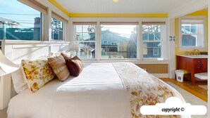 A bedroom with a large bed, a window with a view of houses, a wooden dresser, and a small trash bin. - HOT TUB: ESCAPE MANOR: 600ft to BEACH:  Seaside Vacation Homes, Inc Managed  (Seaside)