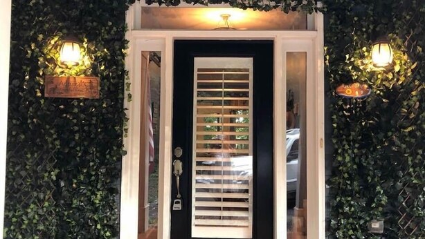 Une porte noire avec une fenêtre, flanquée de deux lampes fixées au mur et d’une enseigne en bois au-dessus de la porte.