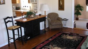 A living room with a wooden floor, a central island with a lamp, two chairs, a sofa, a painting on the wall, and a potted plant.