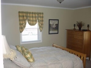 Ein Schlafzimmer mit einem Bett, einer Kommode, einem Fenster mit Vorhängen und einem gerahmten Bild an der Wand.