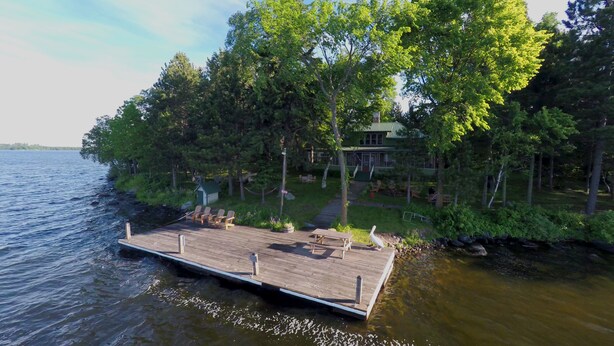 Un muelle de madera se adentra en un lago, rodeado de exuberante vegetación y una casa con tejado verde.