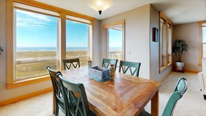 A dining area with a wooden table and chairs, large windows with a view of the beach, and a ceiling light. - OCEAN FRONT: SEAVIEW SERENITY: Seaside Vacation Homes, Inc Managed  (Seaside)