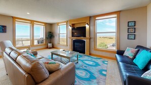 A spacious living room with a leather sofa, a black leather couch, a fireplace, and large windows offering a view of the outdoors. - OCEAN FRONT: SEAVIEW SERENITY: Seaside Vacation Homes, Inc Managed  (Seaside)