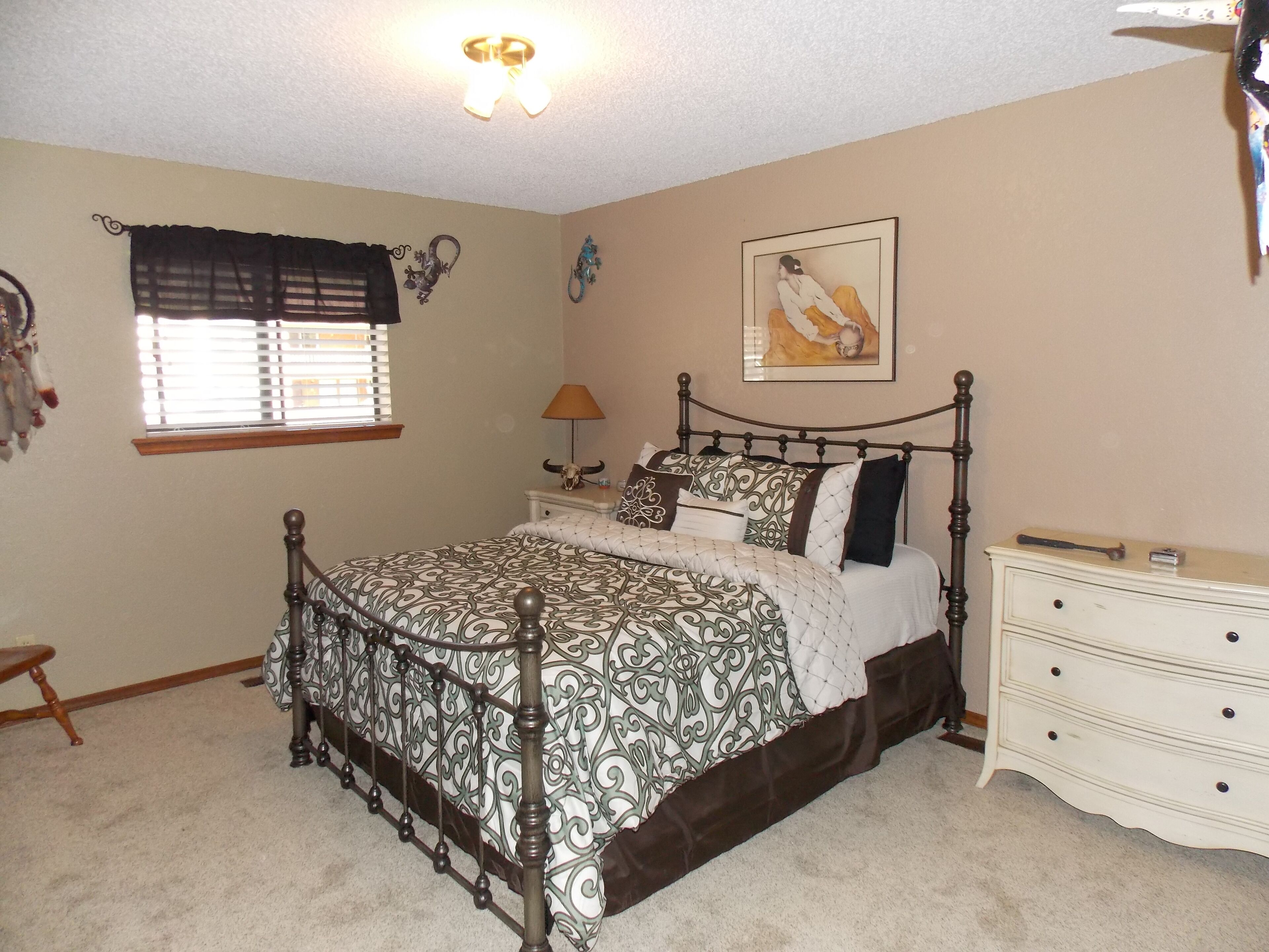 A bedroom with a four-poster bed, a bedside table with a lamp, a dresser, and a wall-mounted painting.