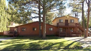 A two-story cabin with a balcony and a deck, surrounded by trees and a grassy yard. - New furniture Beautiful Home! (Ruidoso)
