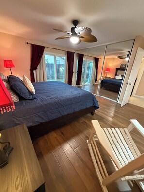 A bedroom with a bed, a ceiling fan, a wooden floor, a mirror, and a view of a backyard.