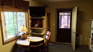 A compact room with a dining table, chairs, a television, and a door leading outside. - Kenai Riverfront Cozy Studio (Soldotna)