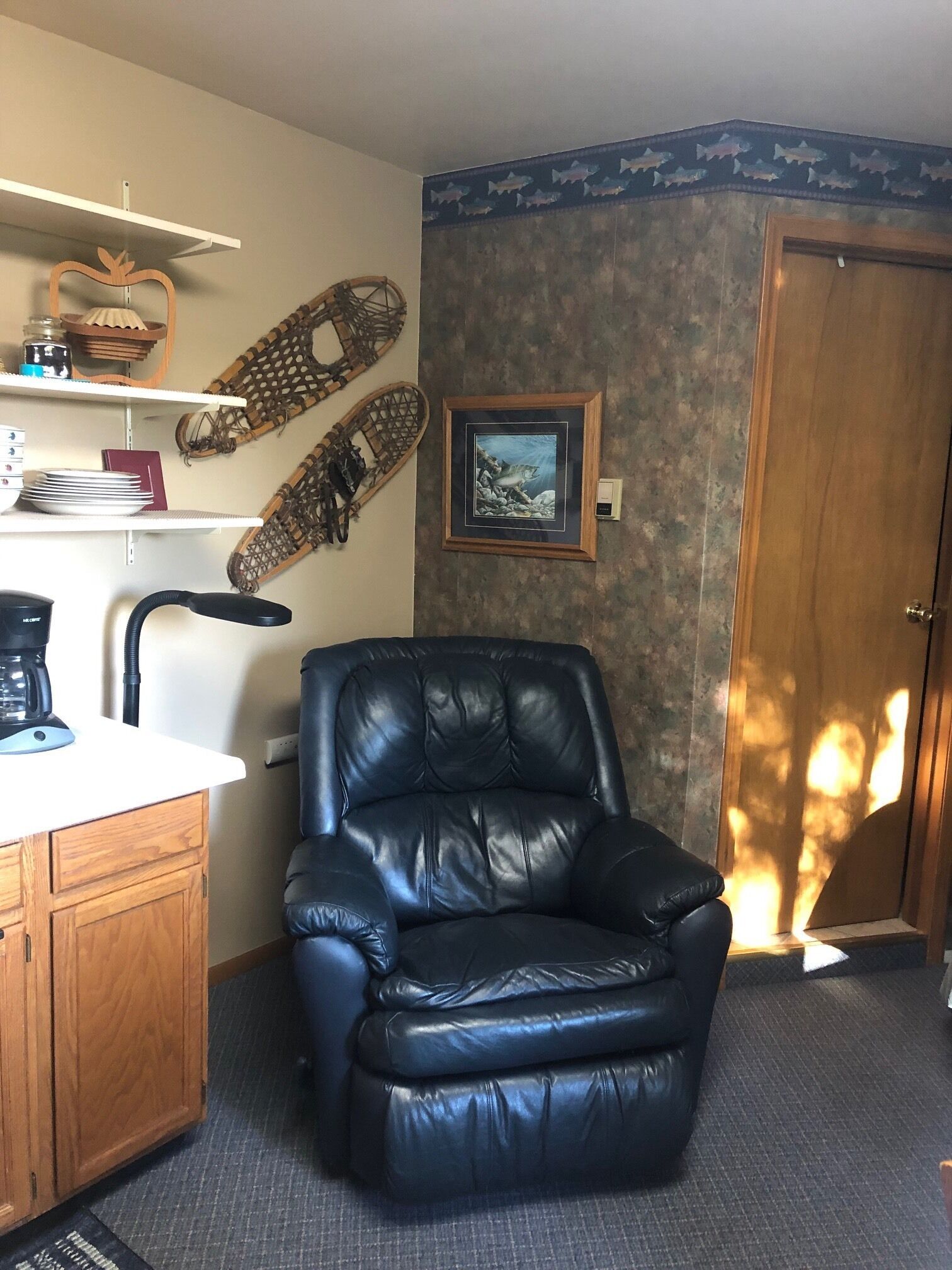 Una habitación con un sillón de cuero negro, una cafetera sobre una encimera blanca, estantes con adornos, un cuadro en la pared y una puerta con marco de madera.