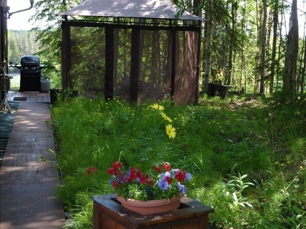 A wooden gazebo with a roof, surrounded by lush greenery and a flower bed with various flowers. - Kenai Riverfront Cozy Studio (Soldotna)
