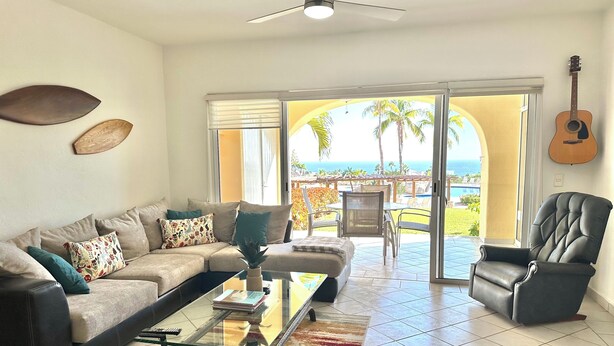 A living room with a sofa, armchair, and coffee table, a guitar on the wall, and a view of the ocean through large glass doors.