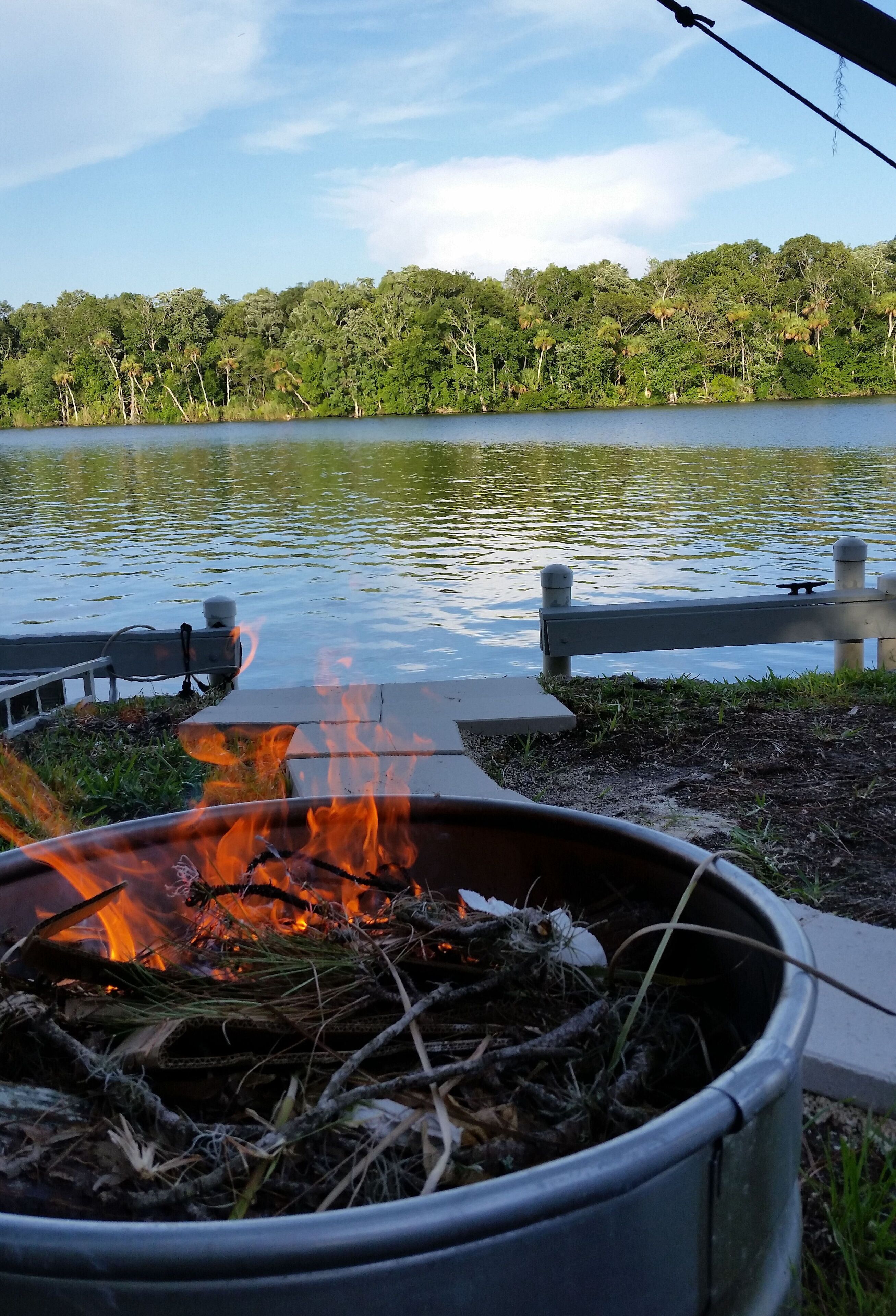 A lakeside view with a fire pit and a bench by the water.
