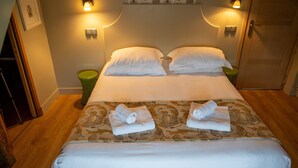 A neatly made bed with white linens and two rolled towels, a wooden floor, and a wall with decorative birdhouse lights.