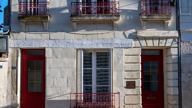 A two-story building with a red door and windows, a balcony with a metal railing, and a chimney.