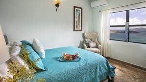 A bedroom with a teal bedspread, a framed picture on the wall, a chair with a cushion, and a tray with fruit and cups. - Ocean-View 3BR Villa for Families and Groups (St Thomas)